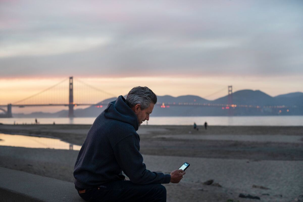 man met grijs haar en donkerblauwe trui gebruikt zijn mobiele telefoon, zittend op het strand van San Francisco